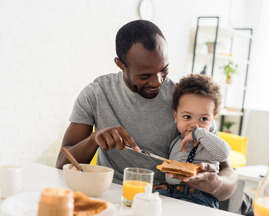 Father with young son on his lap spreading peanut butter on toast.