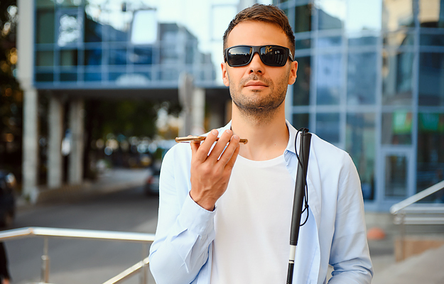 A blind man with sunglasses on holding a cell phone and white cane.