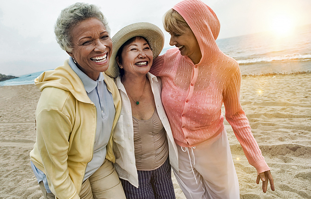 Three mature women smiling and walking on a beach together.