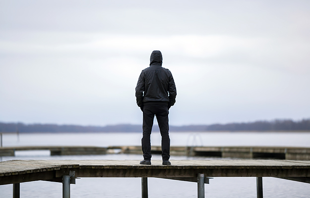 A man standing on a wooden footbridge looking at a lake and overcast winter sky.