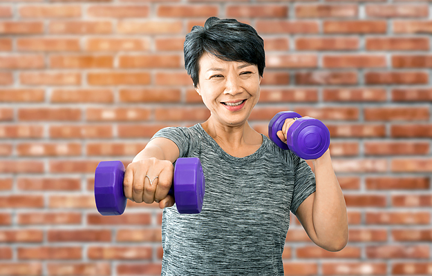 A senior woman working out with dumbbells in front of a brick wall.