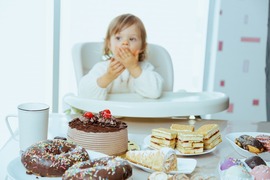 Young toddler in a high chair eating chocolate cake, with an assortment of pastries and cakes on the table in the foreground.