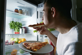 Young man eating pizza from a refrigerator at night.