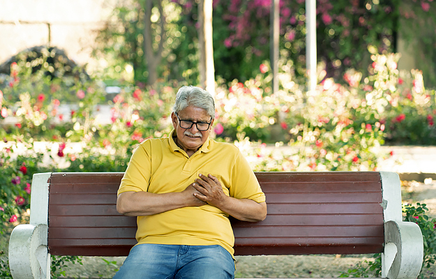 A senior man sitting on a park bench and holding his chest in pain.