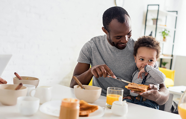 A father with his young son on his lap spreading peanut butter on toast.