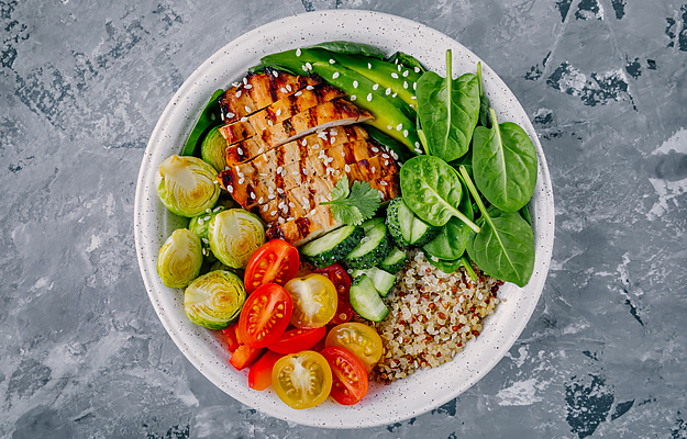 A healthy bowl of food with grilled chicken, avocado, spinach, quinoa, cucumbers, tomatoes, and brussels sprouts on a grey background.