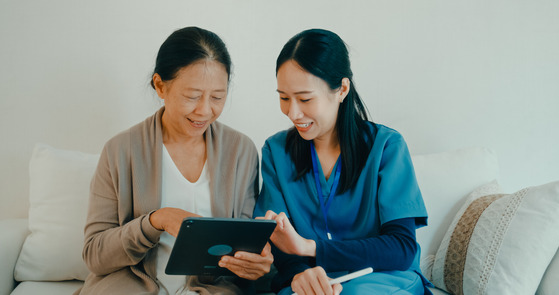 Young Asian nurse uses a tablet to help an elderly woman technology and support on sofa in living room at home.
