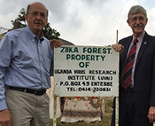 Dr. Francis Collins and Dr. Roger Glass stand in front of the Zika Forest sign in Uganda