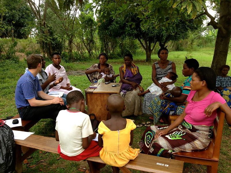 Group of people, consisting of men, women children, sitting outside on wooden benches in a circle and engaging in discussion