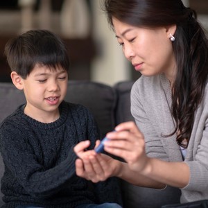 A young Asian boy sits on a sofa beside his mother as she tests his blood sugar level with a lancing device. 