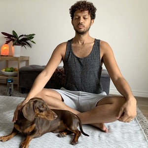 A front view of a mixed race male with his eyes closed meditating with his sausage dog lay down in front of him at home in his living room.