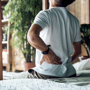 Rear view of senior Asian man suffering from backache, massaging aching muscles while sitting on bed. Elderly and health issues concept