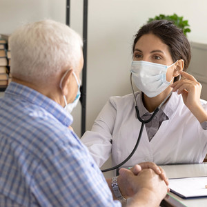 Young female doctor in facemask listen to old male patient heart chest with stethoscope at clinic meeting. 