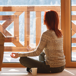 Woman sitting on floor in her home drinking coffee and looking through window at snow covered mountain. She is meditating.