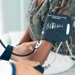 Close up of an older woman sitting on an exam table with a blood pressure cuff on her arm. The doctor is holding the inflation bulb in one hand 