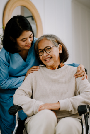An elderly woman patient seated in a wheelchair, being embraced by a nurse from behind.