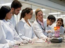 a researcher and students, all in lab coats, standing at a lab bench