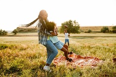 A mother hugging her daughter while spinning her around in an open field while her son tosses a football in the air in the background.