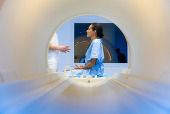 A camera looking through an MRI machine shows a female patient speaking with a health care provider. 