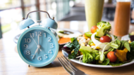 An alarm clock displaying 7 o'clock and a salad on a dining table.