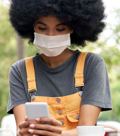 A Black woman wearing a mask sits outside. She is looking down at her cellphone.