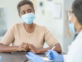A Black woman wearing a blue surgical mask talks to her doctor.
