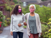 Two women in cloth face masks walk arm in arm.
