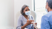 An older woman speaks to her doctor. Both are wearing surgical facemasks.