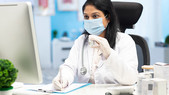 A woman in a surgical mask sits in the lab. She is holding a vial and looking at her computer.