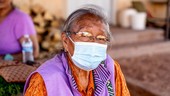 An elderly woman in glasses and a surgical mask looks at the camera. She is wearing brightly colored clothes and sits outside.