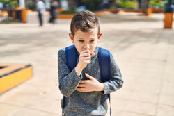 A young boy wearing a backpack covers his mouth and holds his chest while coughing. 