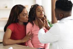 A doctor feels the throat of a young Black patient while she sits on her mother’s lap. 