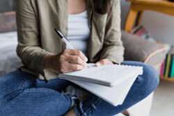 A woman sits with legs crossed writing in a journal. 