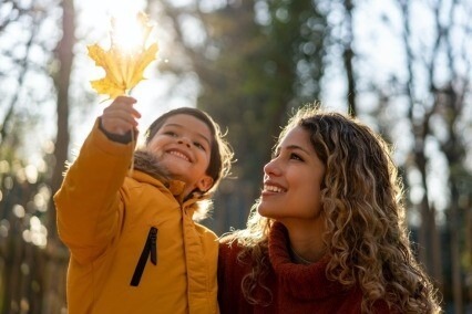 Mother outside on a sunny day with a young child, looking at a yellow maple leaf.