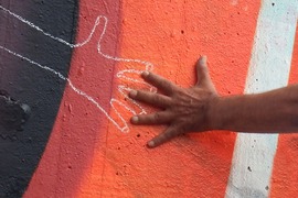 A person's hand touching a chalk outline of another hand on a wall. 