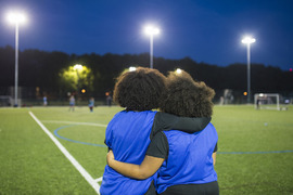 Rear view of two teen girls wearing soccer uniforms and standing outside on an athletic field at dusk, watching a game.