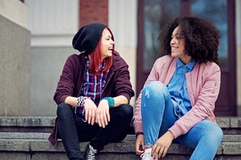 Two teenage girlfriends sitting on steps outdoors and laughing together.