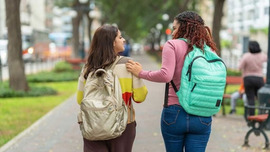 Rear view of a two girls wearing backpacks and walking outside having a friendly conversation together.