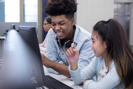 Two teenagers sitting in a classroom and talking to each other in front of a computer screen.