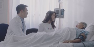 Photo of female Asian patient lying down in hospital bed and speaking with health care provider