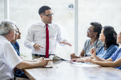 A man standing at the head of the table providing training to a diverse group of colleagues seated at the table.