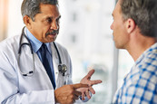 A doctor wearing a white lab coat and stethoscope facing a male patient wearing a white and blue flannel shirt.