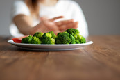 A teen (in background) pushes away a plate with broccoli on it (in foreground).