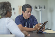 A health care provider sitting with a patient and taking notes on their tablet.