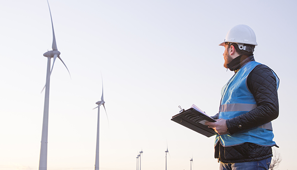 A man in a hard hat is holding a clipboard and looking toward a large wind turbine.