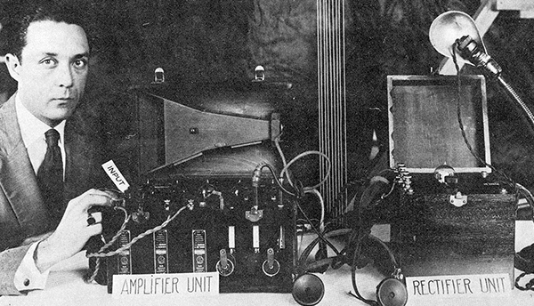 A historical black-and-white photo shows a man in a suit with equipment labeled "Amplifier Unit" and "Rectifier Unit."
