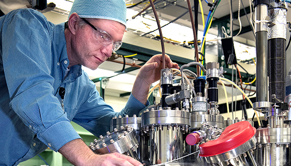 A researcher (Josh Pomeroy) leans over an array of metal equipment (UHV chamber).