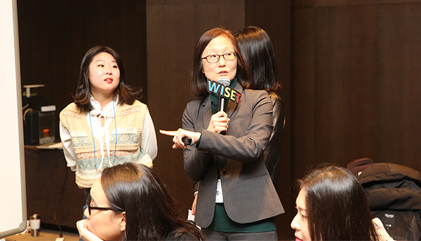 A woman (Hae-Jeong Lee) speaks into a microphone at an event with other women. 