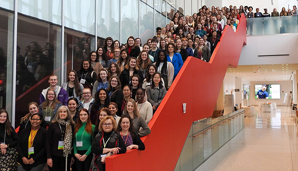 At CUWiP, more than 100 women pose together on a staircase with a bright red wall running down it.