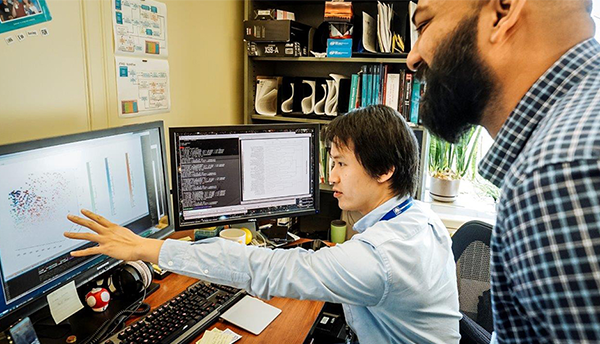 Two men (Tytus Mak and Arun Moorthy) look at a computer screen showing mass spectrometry data.